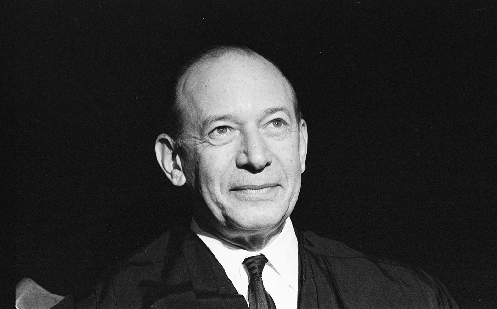 Close-up portrait of a man in a suit and tie, smiling gently, against a dark background, wearing a judicial robe.