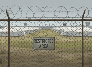 A stark, documentary-style illustration of a federal detention facility seen from outside a chain-link fence topped with razor wire. The scene is overcast and flat. In the foreground, a weathered sign reads "RESTRICTED AREA." In the background, white temporary tent-like structures stretch across the frame. The color palette is muted — grays, yellows, and institutional whites. No people. Bureaucratic, cold, and ambiguous in ownership — could belong to anyone, which is the point. Photorealistic but slightly stylized, editorial illustration style.