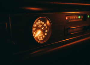 A close-up of a vintage-style AM radio dial glowing amber in the dark interior of a car, the needle resting on a frequency band. Dashboard lights faintly visible in background. Moody, cinematic atmosphere. Documentary photorealistic style, warm amber and deep shadow palette. No text, no logos.