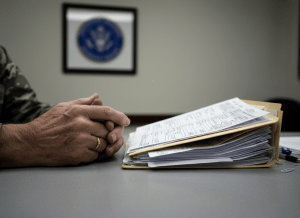 A stark, documentary-style close-up of a veteran's worn hands resting on a plain government desk, next to an open manila folder filled with benefit claim forms, some stamped, some blank. A blurred VA seal is visible on a placard in the soft background. Fluorescent overhead lighting. The aesthetic is institutional and human simultaneously — bureaucratic process meeting real human stakes. Muted tones of beige, pale gray, and olive green. No faces visible. The image evokes quiet administrative failure with serious personal consequences.