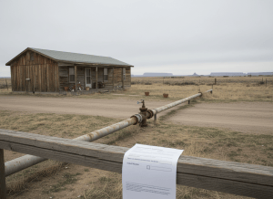 Documentary-style image of a modest home on tribal land — wood-frame, well-worn but clearly lived-in, with a child's bike on the porch and a few potted plants. The water main runs visibly along the road in front of the house, close enough to touch, but does not connect to the home. A government eligibility form lies on the porch railing, partially filled out, with a checkbox next to "Tribal Member" left unchecked. The sky is overcast and the setting is remote, high plains or desert Southwest. The mood is quiet exclusion — not poverty porn, but the specific, structural kind of being left just outside a line. Muted, dry, institutional aesthetic.