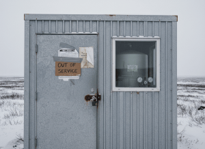 Documentary-style close-up of a rural Alaska water treatment facility — a small, utilitarian building in a remote village setting, snow-dusted tundra visible in the background. The door has a padlock on it and a handwritten sign reading "Out of Service." Beside the door, a public notice is tacked to the wall — partially legible, with the words "Boil Water Advisory" visible. A chlorination tank is visible through a small window, its gauges unmonitored. The facility looks functional but unattended. The mood is quiet public health emergency — not dramatic, but the kind of systemic neglect that accumulates until something breaks. Muted, cold, institutional aesthetic — gray, pale blue, weathered aluminum.