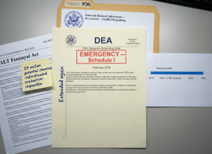 Documentary-style flat-lay on a federal regulatory desk. Center: a DEA temporary scheduling order stamped "EMERGENCY — Schedule I" dated February 2018, with a handwritten note in the margin reading "Extended again." Next to it, a printed timeline on graph paper showing a horizontal bar labeled "Temporary Order" stretching from 2018 to 2025 — seven years — with tick marks at the original two-year endpoint. To the left, a Congressional record page showing the HALT Fentanyl Act, dated July 2025. A sticky note reads "24 million potential chemicals — individualized evaluation: impossible." In the background, an FDA evaluation folder labeled "Fentanyl Related Substances — 46 reviewed / ~24,000,000 pending." The mood is bureaucratic inadequacy confronted by chemistry it cannot pace — not dramatic, procedurally damning. Muted institutional aesthetic — gray, cream, DEA seal blue, fluorescent light.