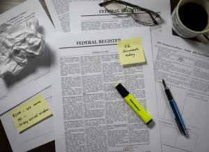 Documentary-style flat-lay on a federal policy analyst's desk. Center: a printed copy of the Federal Register dated September 24, 2026, open to a dense page of rule notices. Visible headlines on the page: "NRC: Wholesale Revision of Reactor Licensing Rules," "Commerce: Polysilicon Import Restriction — Effective Retroactively," "FTC: Post-Employment Ethics Screen Eliminated." A sticky note reads: "106 documents today." Scattered nearby: a printout labeled "Confirmatory Order — Dept. of the Air Force — Kirtland AFB Radioactive Source Event"; a yellow highlighter resting on the phrase "temporary final rule — no prior notice"; a small notecard reading "$20M — sole source — IRC — 30-day arrival window." The mood is the daily grind of federal rulemaking, dense and unglamorous. Muted palette — newsprint gray, federal blue, highlighter yellow, government cream, red pen marks.